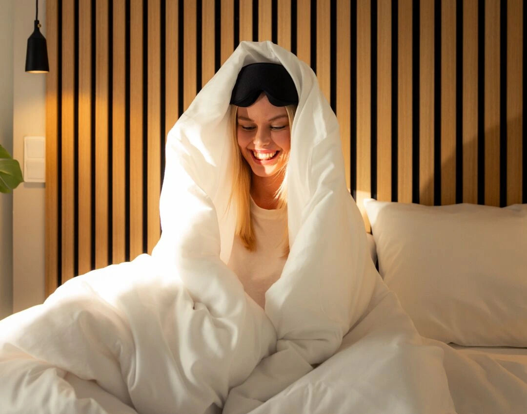 Woman in bed under a white duvet with a sleep mask on, smiling.